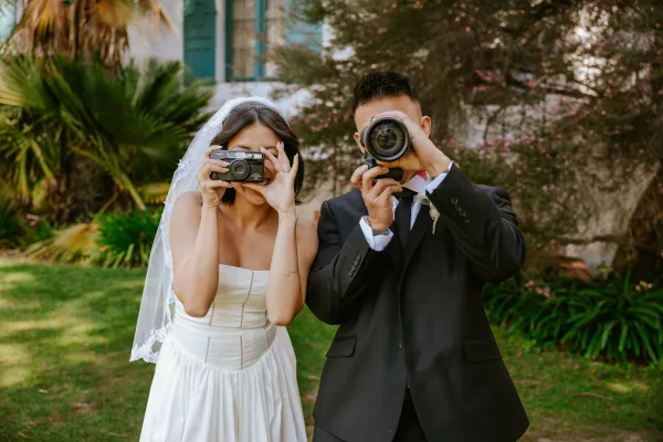 Couple portrait of bride and groom with cameras, bride in strapless dress and veil showing ring on a garden lawn with palms