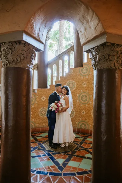 Couple portrait of bride and groom smiling as he kisses her cheek, bride holding a bouquet under an arched doorway with stone columns