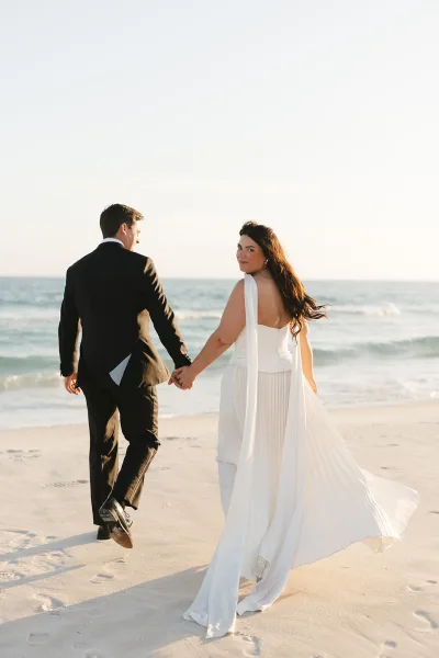 Couple portrait of bride and groom walking hand in hand along a sandy beach, with ocean waves rolling in and ribbons fluttering from her gown.