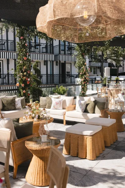 Wedding lounge setup for outdoor cocktail hour seating with rattan sofas beside a pool, framed by hotel balconies and striped awnings.