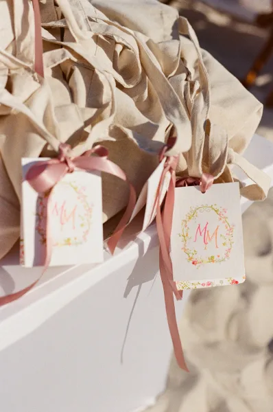 Wedding welcome bags in a canvas welcome tote tied with satin ribbon, arranged on beach sand to greet arriving guests.