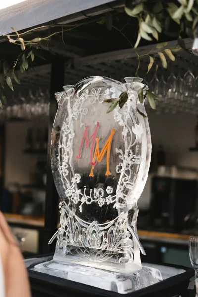 Wedding ice sculpture, a sleek monogram ice sculpture, centered on a bar counter beneath hanging wine glasses, with subtle greenery.