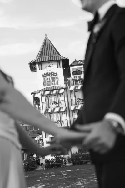 Couple portrait of the bride and groom holding hands beneath a clock tower, his bow tie neat, standing on a lawn with striped awnings.