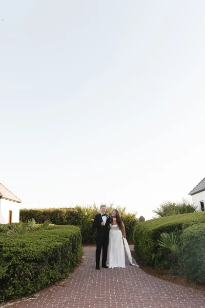 Couple portrait of a bride and groom laughing, arm in arm, the groom in a black tuxedo, on a brick path by hedges and white buildings under blue sky.
