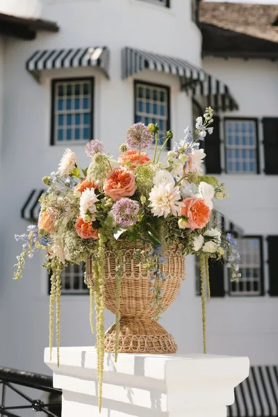 Wedding flower arrangement of ceremony urn flowers overflowing from a wicker urn, set before a white building facade with striped awnings.