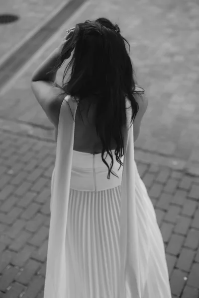 Wedding dress detail of an open back wedding dress, its pleated skirt trailing over brick pavement on a quiet street.