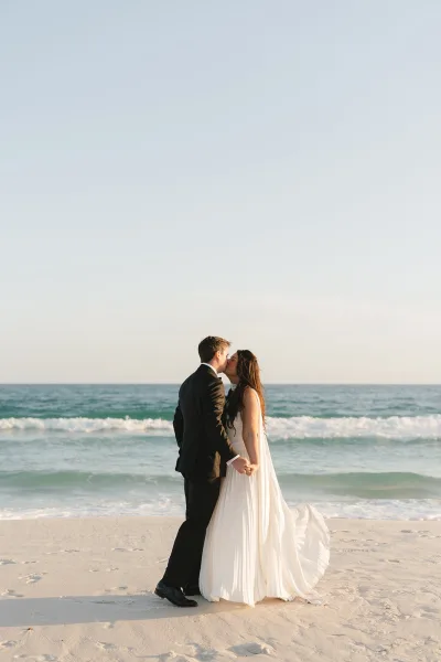 Wedding kiss portrait of a bride and groom on a sandy shore, a beach wedding portrait framed by rolling ocean waves and a sweeping dress train.