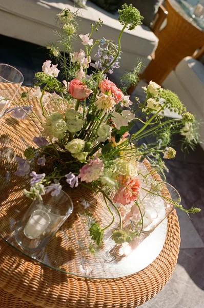 Wedding centerpiece, a pastel garden centerpiece of peach and blush blooms on a rattan table, with sofa seating and a wicker chair behind.