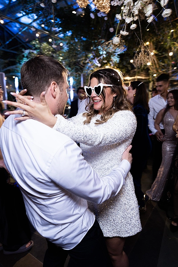 Wedding reception dancing as bride in heart-shaped sunglasses and sequined mini dress hugs groom under string lights in a greenhouse venue