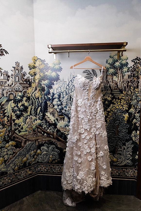 Wedding dress with lace bridal gown details and floral appliqué hanging from a wooden hanger on a brass rack against mural wallpaper
