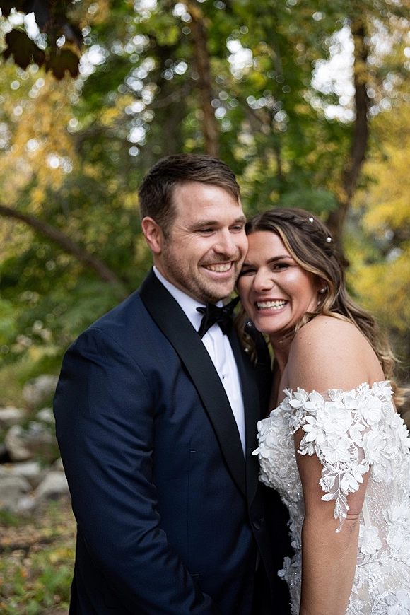 Couple portrait of bride and groom hugging, smiling in natural light, with lace wedding dress and black tuxedo amid green trees