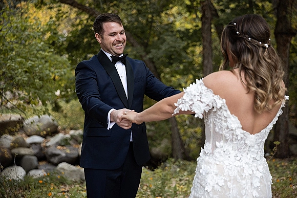 First look moment as bride in an off shoulder wedding dress and groom in tuxedo hold hands by a forest creek with rocks and trees