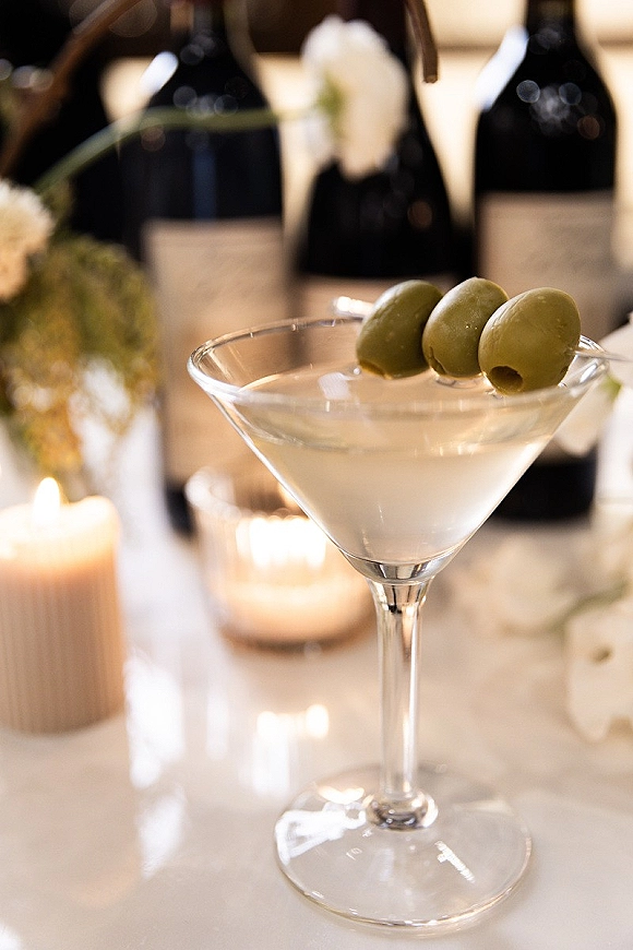 Wedding signature cocktail in a martini glass with olive garnish beside wine bottles, candles, and white flowers on a softly lit reception table
