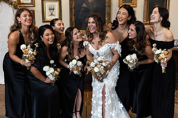Bridesmaid group portrait with bride with bridesmaids laughing, holding bouquets in a vintage room with framed paintings and brick floor