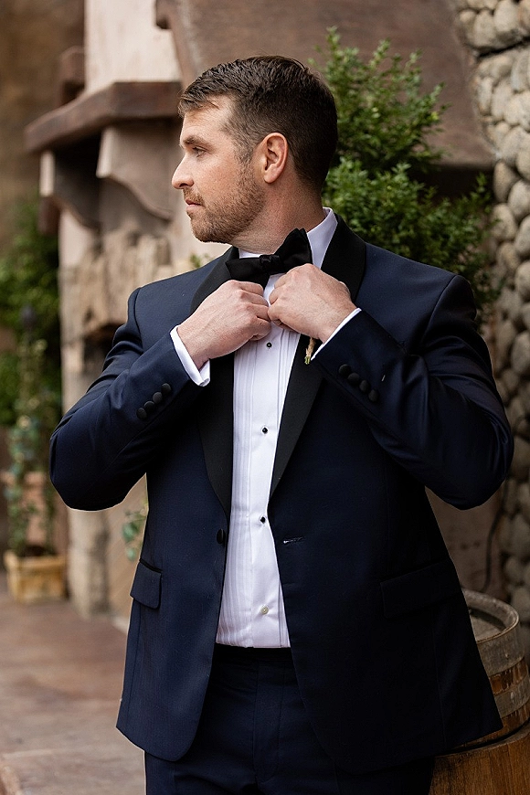 Groom portrait of a man adjusting his bow tie in a classic black tuxedo with boutonniere on an outdoor patio by a stone wall