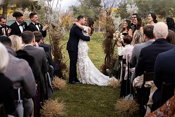 Ceremony kiss as bride in lace gown with long train kisses groom in tuxedo on an outdoor aisle with pampas grass and mountains