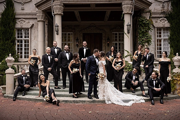 Wedding party portrait of bride and groom with wedding party on stone manor steps, bride in off-shoulder gown holding bouquet, all laughing