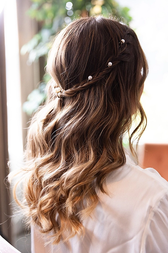 Bridal hairstyle with half up half down wedding hair styled in loose waves, braided crown braid, and pearl hair pins in window light with greenery