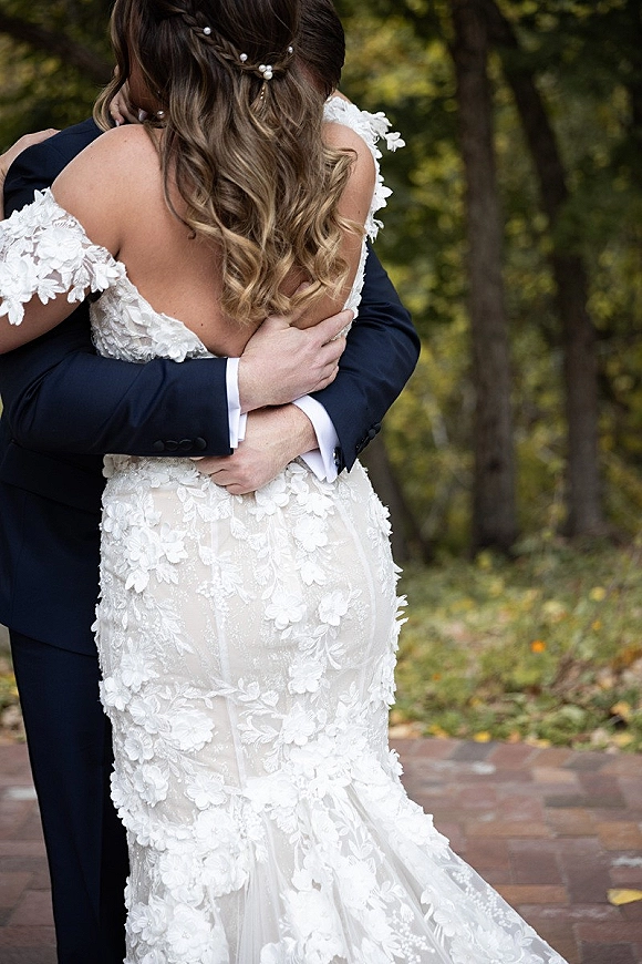 Wedding couple embrace as the groom hugs the bride from behind, highlighting her lace off-the-shoulder gown and pearl pins on a tree-lined path