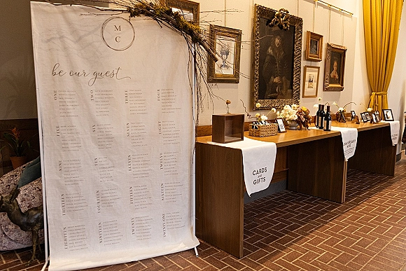 Wedding welcome table with wedding seating chart sign, calligraphy decor, wine bottles, framed photos, and branches against a gallery wall