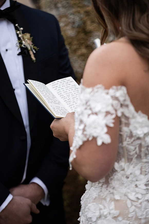 Wedding vows as bride in off the shoulder lace dress reads handwritten wedding vows from a vow book, groom in tux listens by stone wall