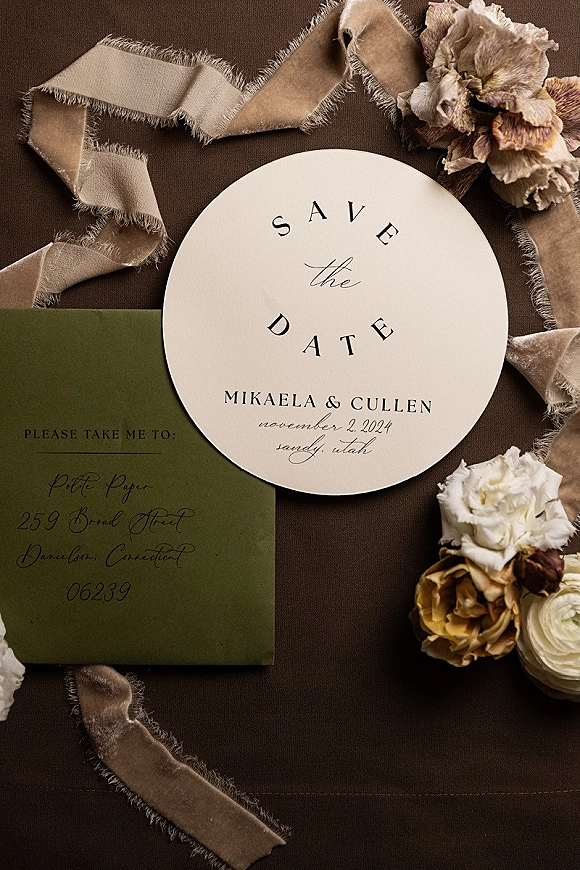 Save the date card with a round invitation and olive green envelope, ribbon and wax seal on a brown fabric backdrop with pressed flowers