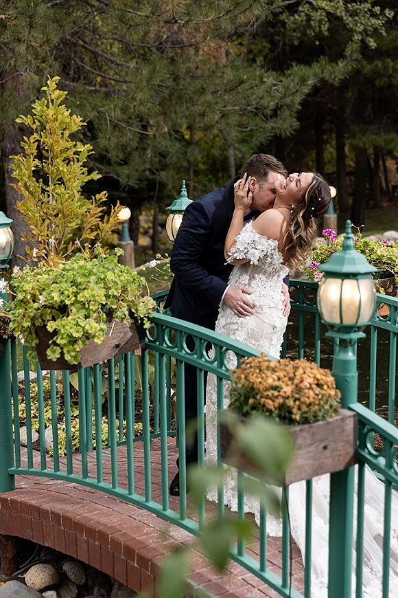 Wedding kiss portrait of bride and groom kissing on a garden bridge, bride laughing in off-the-shoulder gown, lantern lights behind