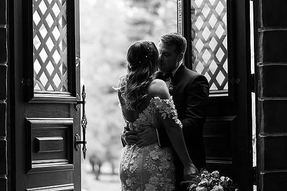 Wedding kiss as the bride and groom kissing in a stone doorway, groom holding her close as she clutches a bouquet, lace off-shoulder gown visible