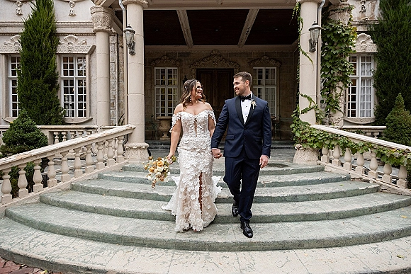 Couple portrait of bride and groom holding hands, bride in off the shoulder lace dress with bouquet on an ivy-lined mansion staircase.