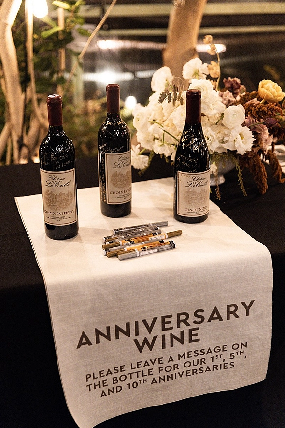 Anniversary wine table with wedding anniversary wine bottles set for guests to sign using metallic markers on a white linen cloth under string lights