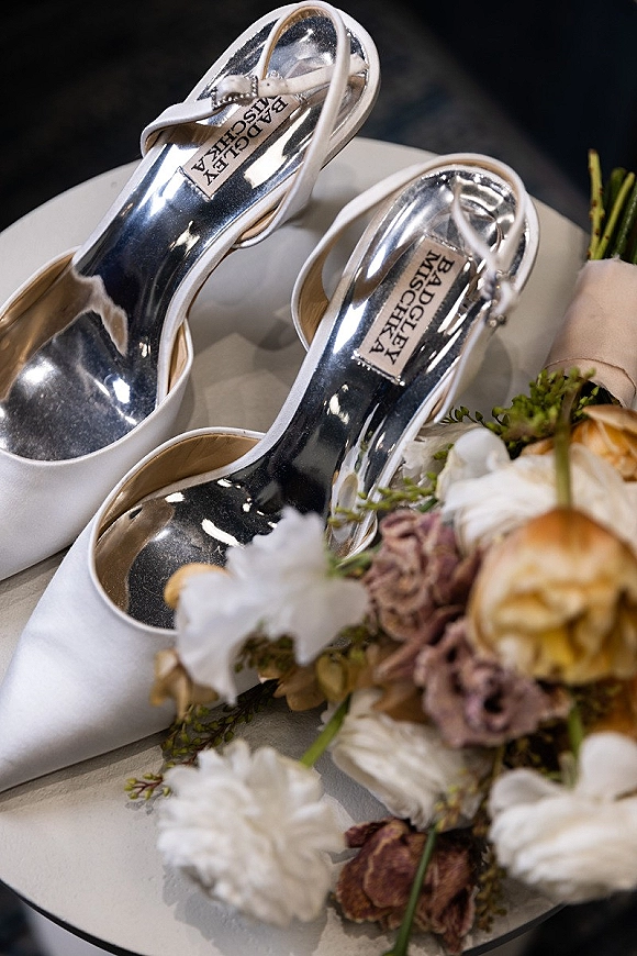 Bridal shoes, white wedding heels with metallic insoles beside a rose and carnation bouquet with greenery on a tabletop against a dark backdrop