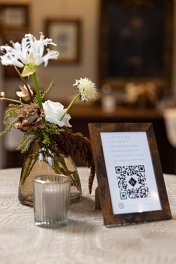 Wedding QR sign with a wedding photo upload QR code in a wood frame on linen, beside a bud vase and glass votive in warm indoor light