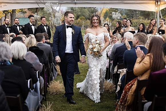 Wedding recessional as bride and groom walk the aisle holding hands, bouquet raised, under a tent canopy with guests and pampas grass decor