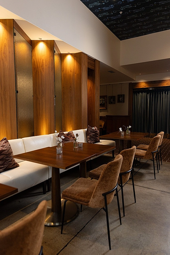 Reception lounge seating with warm wood dining tables, upholstered banquette and chairs, accented by bud vases and votive candles under pendant lights