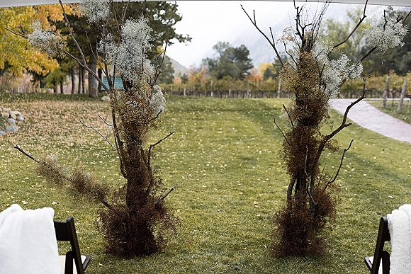 Ceremony altar with branch wedding altar and baby's breath decor, flanked by black chairs on a leaf-strewn lawn with mountains beyond