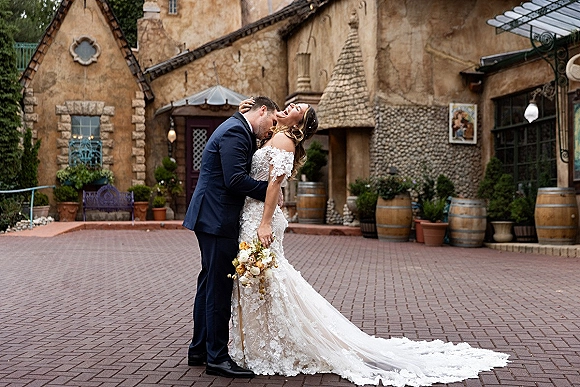 Wedding couple portrait of groom in a navy suit kissing bride’s forehead as she leans back holding a yellow bouquet in a courtyard by stone building and barrels