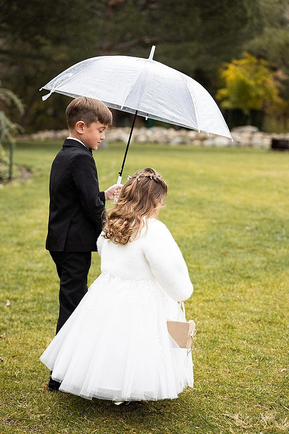 Flower girl and ring bearer with a clear umbrella, her in a white tulle dress and fur wrap, on a grassy lawn by a stone wall