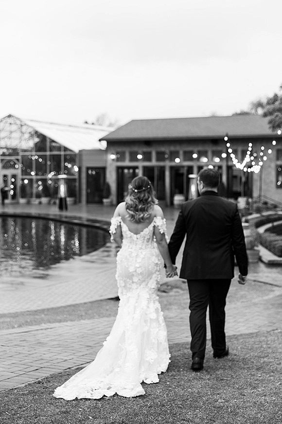 Couple portrait of newlyweds holding hands, walking away by a pool at dusk, bride’s lace gown train flowing under string lights