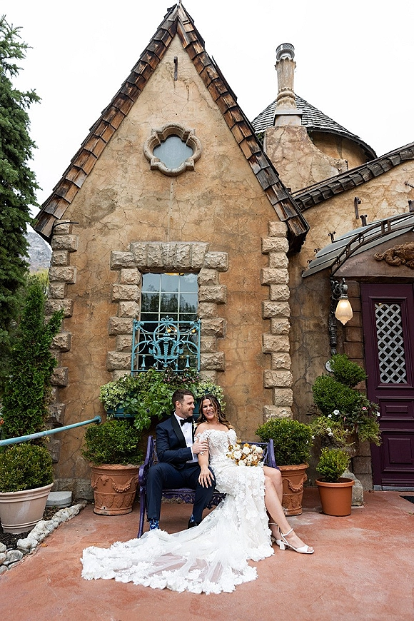 Couple portrait of bride and groom sitting on a bench, bride holding a bouquet, against a stucco villa courtyard facade with potted plants