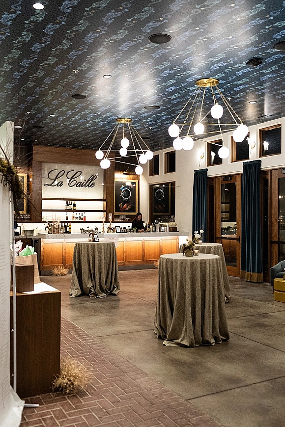 Cocktail hour setup with wedding cocktail tables, draped linens, floral centerpieces and votive candles beside a bar under globe chandeliers