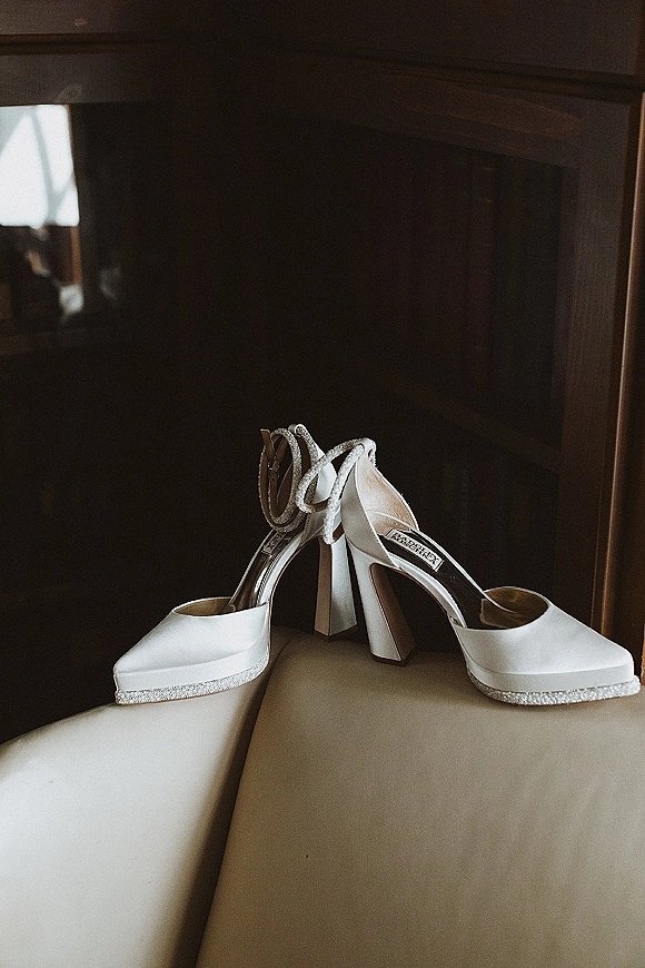 Wedding shoes, white high heel sandals with ankle straps and rhinestone trim, resting on a leather chair in window light with dark wood paneling