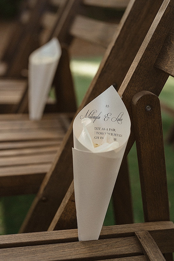 Petal toss cones with flower petals and a printed instruction card attached to wooden folding chairs along a grassy ceremony aisle