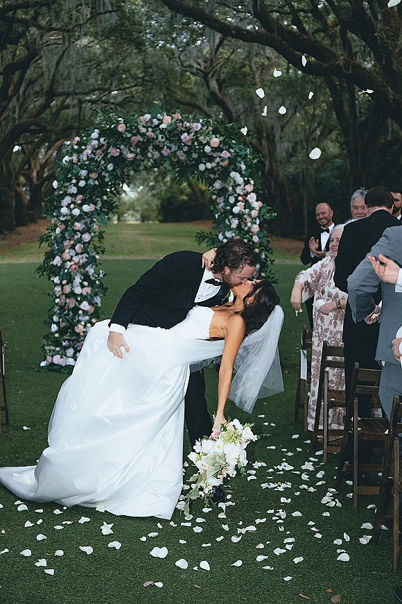 Wedding kiss portrait of bride and groom in a dip kiss wedding photo beneath a floral arch, rose petals on the aisle, guests under mossy trees
