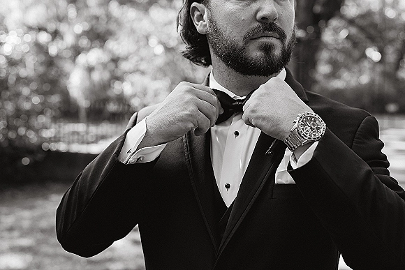 Groom portrait with groom adjusting bow tie, wearing a black tuxedo with cufflinks and wristwatch, standing before blurred trees and bokeh lights