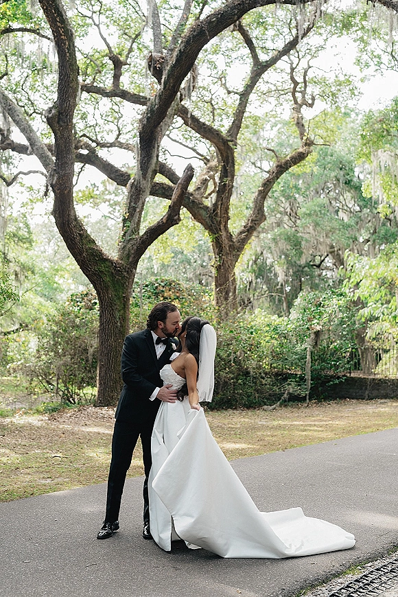 Wedding kiss portrait of bride and groom kissing, her long veil and train flowing as they stand on a garden path under mossy oaks