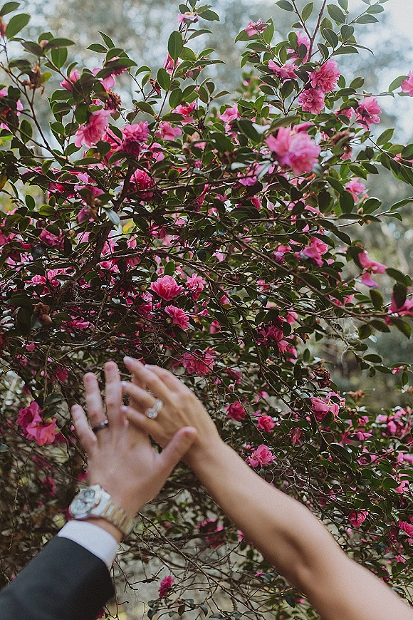 Wedding ring photo of intertwined hands showing an engagement ring and band beside a wristwatch, framed by pink flowers and green leaves outdoors