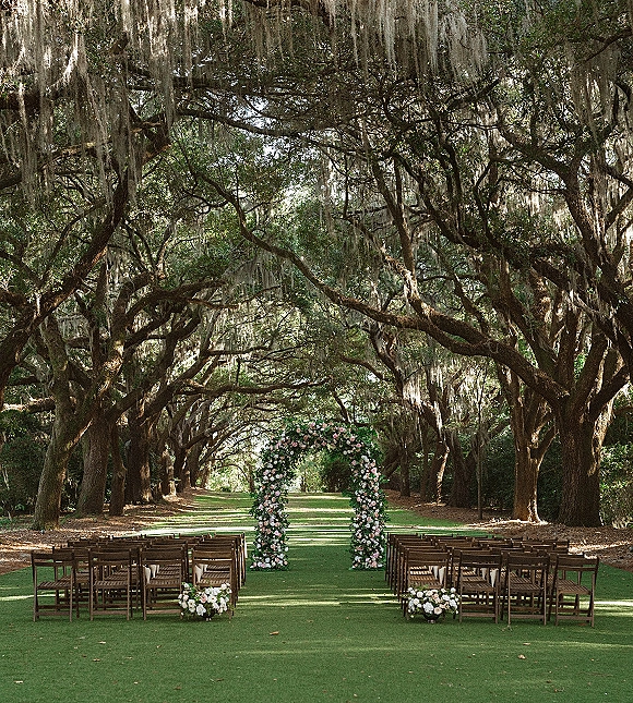 Ceremony setup for outdoor wedding ceremony with a floral arch of pink and white flowers, wooden chairs, and a green runner beneath oak trees with hanging moss