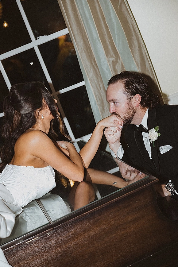 Couple portrait of groom kissing bride’s hand by a window, her strapless dress and his black tuxedo lit softly against night sky outside