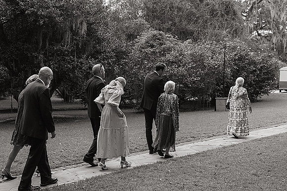 Wedding guests walking in formal suits and midi dresses, one with a shawl, along a stone garden path under mossy trees and string lights