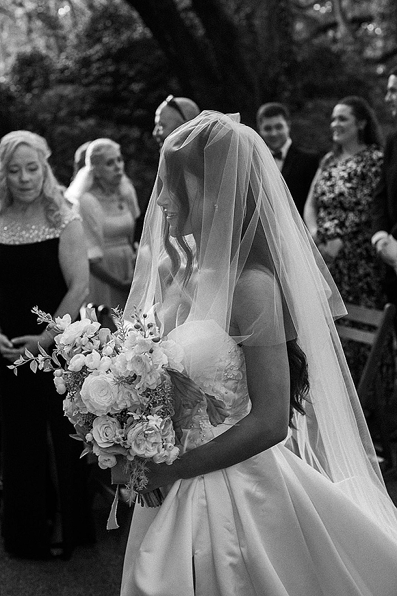 Bridal portrait of a bride holding bouquet with a long veil in side profile, walking the garden ceremony aisle past guests and chairs
